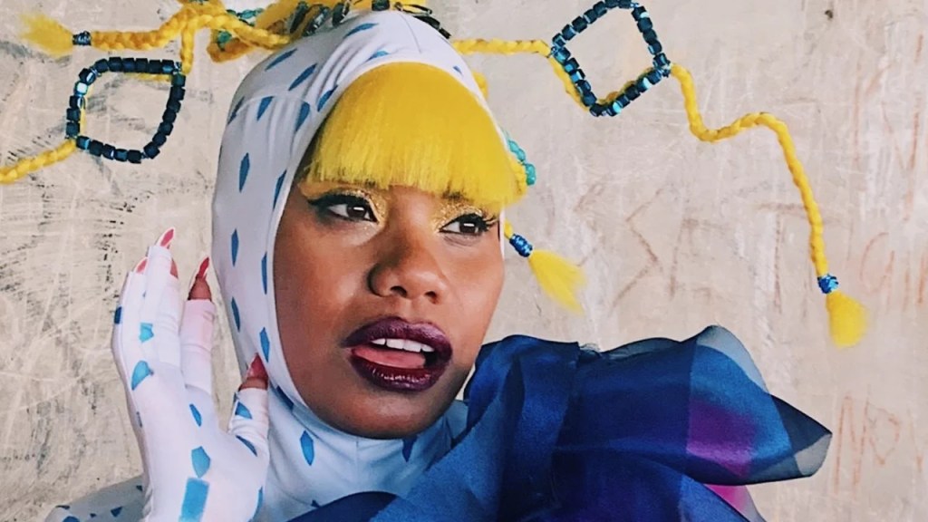 Closeup of Ngaiire wearing a white bodysuit with blue shapes, pink nails, and an intricate head piece prominently featuring yellow hair braided and sculpted with blue diamonds in the design. 